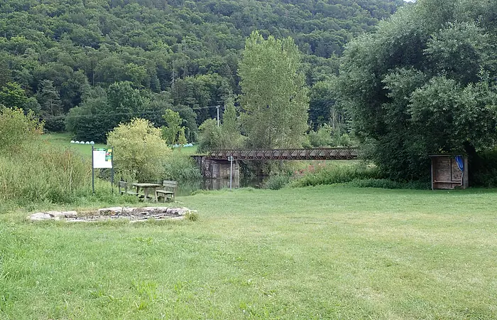 Grüne Wiese mit Feuerstelle, Holztisch und Bänken, Brücke über Fluss und bewaldeter Hügel im Hintergrund.