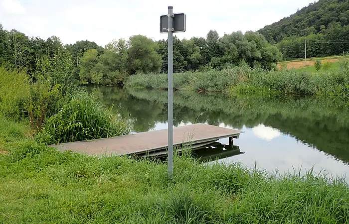 Steg an einem ruhigen Fluss mit grünem Ufer und einem Schildpfosten im Vordergrund.