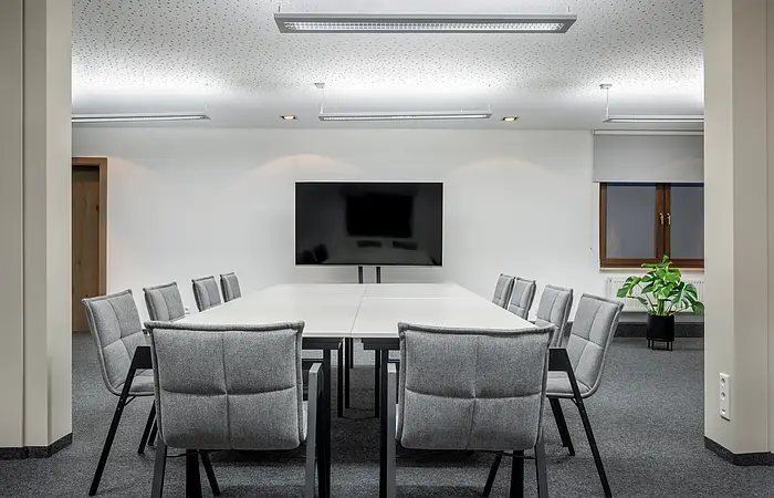 Konferenzraum mit weißem Tisch, zwölf grauen Stühlen, großem Bildschirm und Pflanze am Fenster.