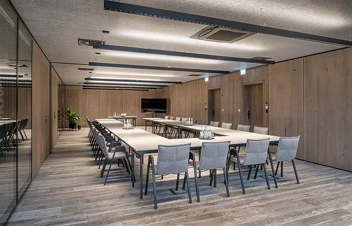 Konferenzraum mit U-förmigen Tischen, grauen Stühlen, Holzwänden und Deckenleuchten.