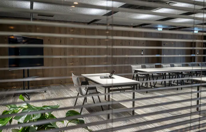 Blick durch Jalousien auf leeren Konferenzraum mit Tischen, Stühlen und offenem Laptop auf Tisch.