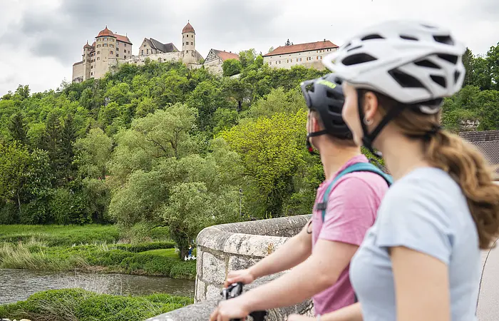 Zwei Personen mit Fahrrädern und Helmen stehen auf einer Brücke und blicken auf eine Burg auf einem Hügel.