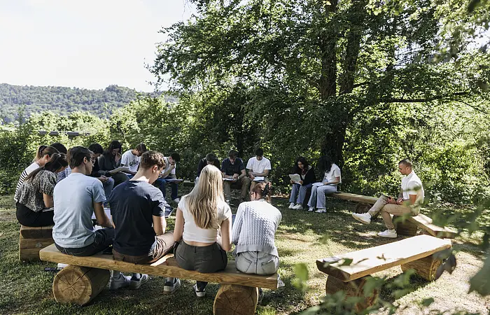 Gruppe von Menschen sitzt im Kreis auf Holzbänken im Freien und liest oder schreibt.