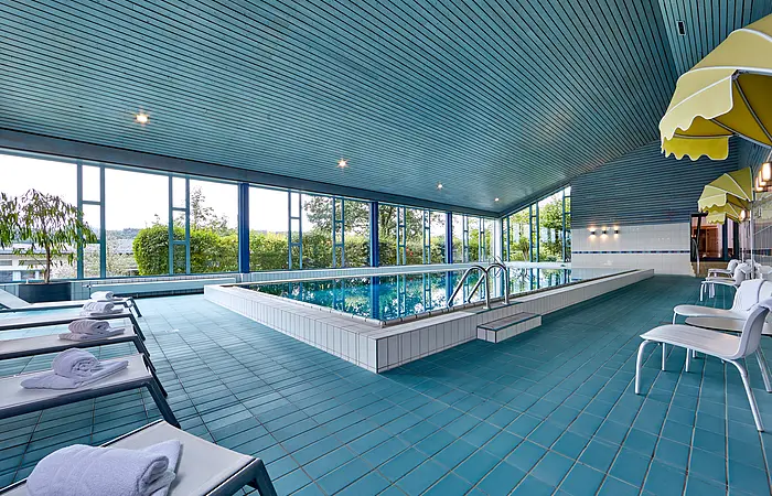 Innenaufnahme eines Schwimmbads mit blauen Fliesen, Liegestühlen mit weißen Handtüchern und großen Fenstern zur Außenbegrünung.