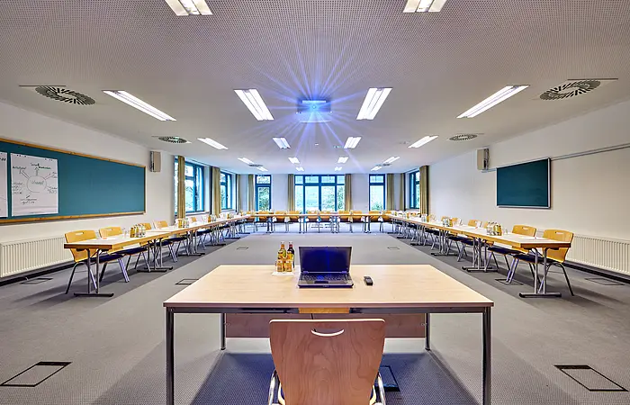 Konferenzraum mit U-förmiger Tischanordnung, Stühlen, Laptop und Getränken auf dem vorderen Tisch, Leinwand an der Decke.