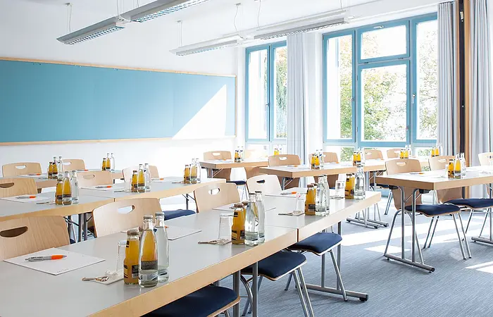 Konferenzraum mit Tischen, Stühlen, Wasserflaschen, Saftflaschen, Gläsern und Schreibmaterial vor großen Fenstern.