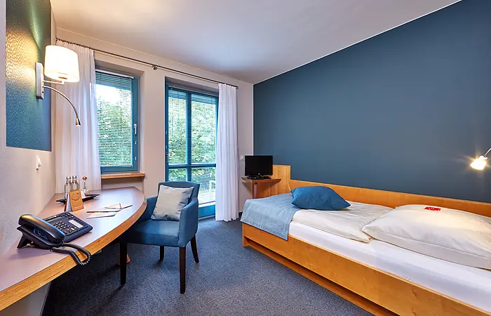 Hotelzimmer mit Einzelbett, Schreibtisch, Stuhl, Telefon, Fenster mit Vorhängen und Fernseher auf kleinem Tisch.
