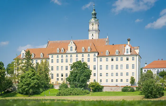 Großes, mehrstöckiges Gebäude mit orangefarbenem Dach und Turm, umgeben von Bäumen und Wiese, unter blauem Himmel.