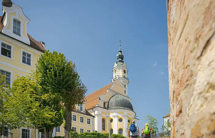 Zwei Personen mit Rucksäcken gehen auf eine gelbe Kirche mit Turm zu, umgeben von Bäumen.