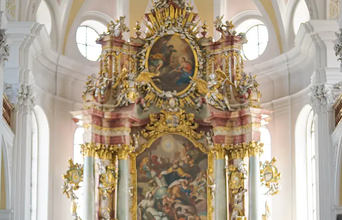Zwei Personen mit Rucksäcken stehen in einer Kirche vor einem reich verzierten Altar.