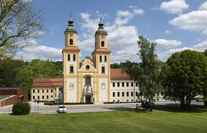 Das Bild zeigt die Klosterkirche St. Johannes der Täufer in Frontalansicht. Zwei barock gehaltene Kirchtürme fassen links und rechts das Mittelschiff und damit auch den Eingang der Kirche. Die Türme sind mehrgeschossig und haben ein Haubendach als Abschluss.  An der rechten Kirchenseite schließt ein langgezogenener Konventbau an. Alle Gebäudeteile sind in ein einem warmen Gelb- und Weißton gehalten. Den Hintergrund des Bildes bildet der langgezogene Bergrücken des Frauenberges.