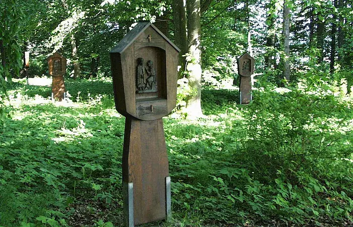 Holztafeln auf einem Kreuzweg mit religiösen Schnitzereien in einem bewaldeten Gebiet.