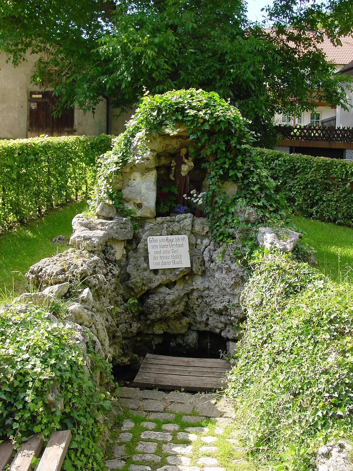 Antoniusbrunnen Im Mittelpunkt der Antoniusbrunnen umgeben von grünem Bewuchs im Hintergrund sind Häuser zu erkennnen.