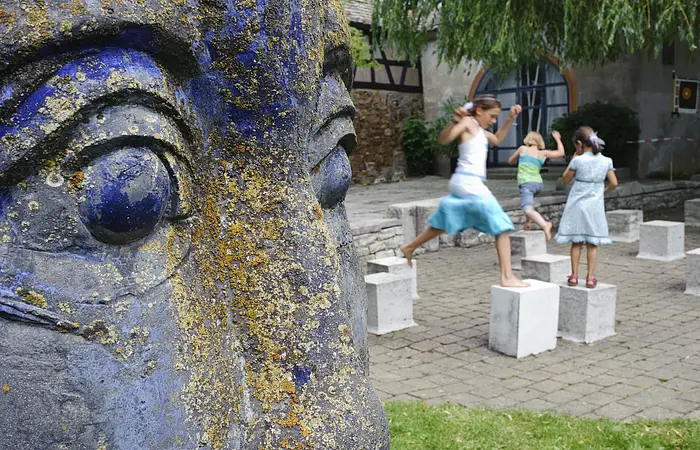 Rechts im Bild die Nahaufnahme des Gesichts einer blauen Statue. Dahinter stehen Steinwürfel in einem Hof. Drei junge Mädchen hüpfen von einem Würfel zum anderen.