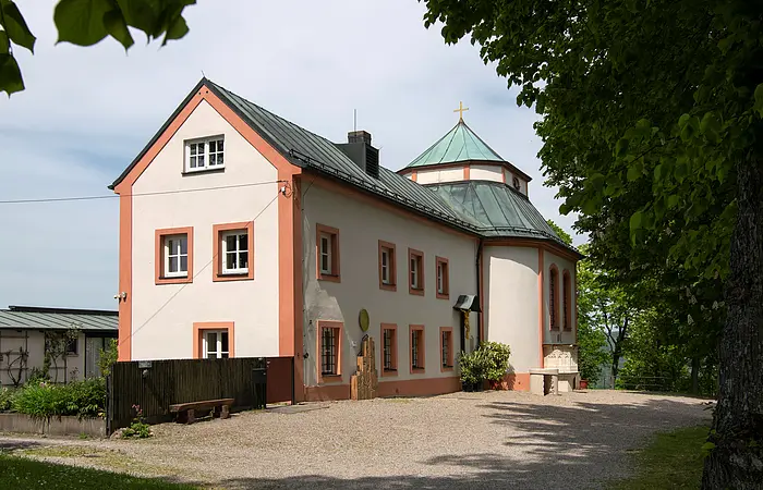 In der Mitte des Bildes steht die Frauenbergkapelle mit angebautem Mesnerhaus. Das Mesnerhaus mit vielen Fenstern und einem Holzkreuz steht dabei im Vordergrund, die schlichte kleine Kapelle im Hintergrund. An der Kapellenseite sind zwei große Fenster und eine Kanzel aus Stein. Das Kapellendach hat eine geknickte Dachfläche mit steilerem Unterdach und flacherem Oberdach. An der Spitze ist ein schlichtes goldenes Kreuz angebracht. Sowohl das Haus, als auch die Kapelle sind cremefarben mit lachsfarbenen Hauskanten und Lisenen. Der Vorplatz ist mit grauem Schotter bedeckt.