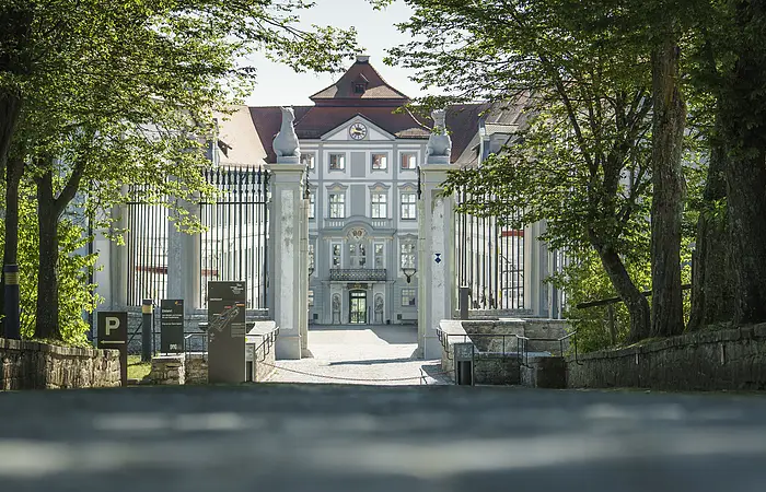 Eingangstor zu einem historischen Gebäude mit Bäumen und Schildern am Wegesrand bei Tageslicht.