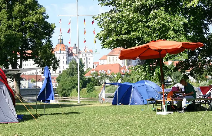Der Platz mit einigen Zelten und einem orangefarbenem Sonnenschirm. Durch zwei Bäume sieht man das Schloß Neuburg.