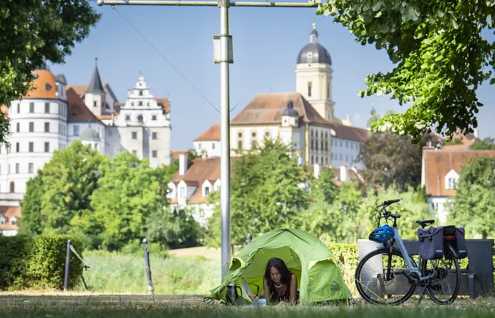 Eine Frau in einem grünen Ein-Mann-Zelt schaut heraus. Rechts neben ihr steht ein Fahrrad. Das Schloss thront hinter ihr.