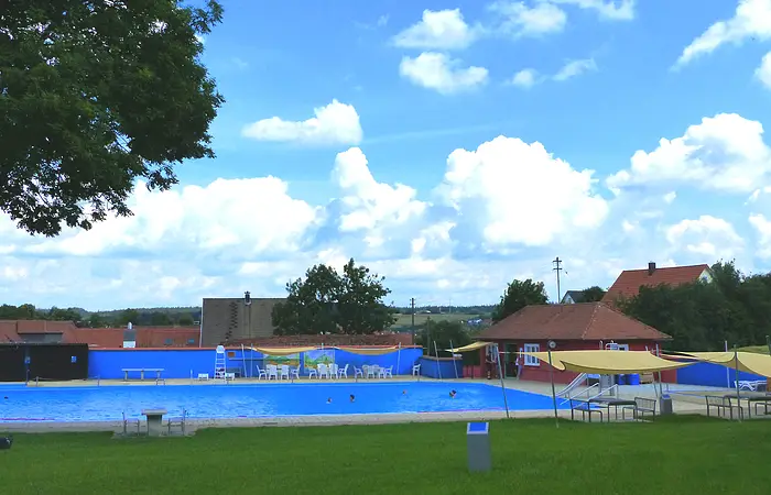 Freibad mit blauem Wasser, umgeben von Rasen, Bäumen und Gebäuden unter einem bewölkten Himmel. Vorderhalb vom Becken sind Sitzmöglichkeiten.