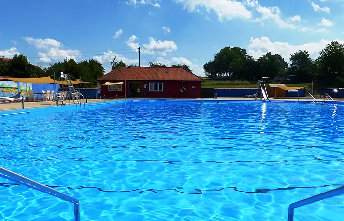 Das Freibad in Monheim: Schwimmbecken mit klarem Wasser, umgeben von Liegestühlen, einem roten Gebäude und Umkleidekabinen im Hintergrund.