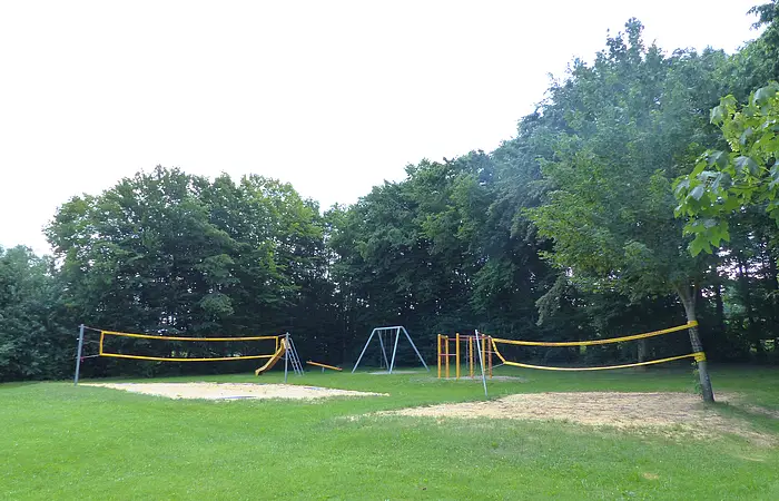 Spielplatz mit zwei gelben Volleyballnetzen, einer Rutsche, einer Schaukel und Klettergerüst vor Baumreihe auf Grasfläche, bei Tageslicht.