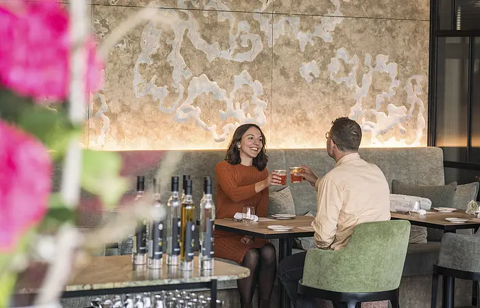 Zwei Personen sitzen in einem Restaurant und stoßen mit Getränken an, im Vordergrund unscharfe Blumen und Flaschen.