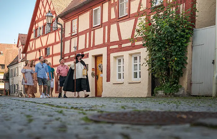 Vorbei an einem renovierten Fachwerkhaus kommt eine Gruppe Menschen auf den Betrachter zu, vorne ein Mann in altertümlicher Kleidung mit großem Hut, Umhang und Laterne.