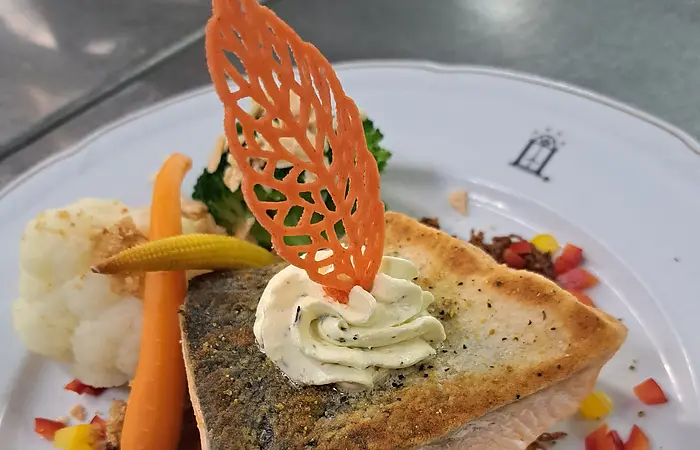 Ein rosa gebratenes Stück Lacks mit einem Blatt und Merrettich dekoriert, liegt auf einem Kartoffelsrösti. Das weiße Teller ist zudem mit Blumenkohl, Karotte und Paprika dekoriert.
