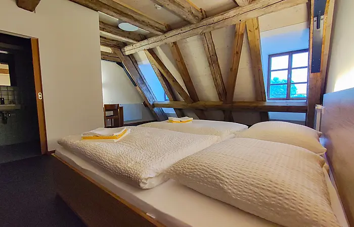 Schlafzimmer mit Dachschräge, Holzbalken, zwei Betten mit weißen Decken und Kissen, Fenster und offenem Badezimmer.