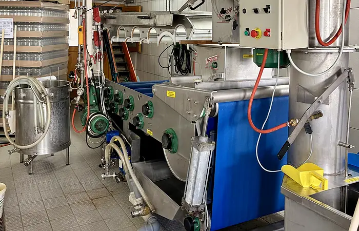 Industrielle Maschine mit Steuerung zum Entsaften von Obst. Rohre und Förderband in einem Raum mit Fliesenboden und -wänden.