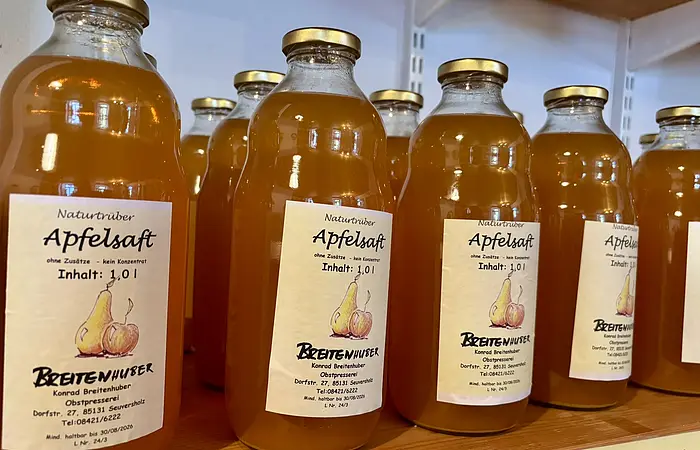 Mehrere Flaschen naturtrüber Apfelsaft mit goldfarbenen Deckeln auf einem Holzregal.