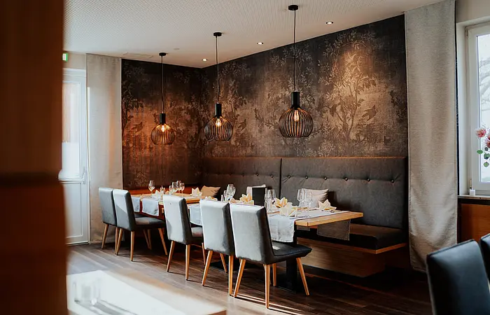 Es ist eine lange Tafel mit gepolsterten Stühlen und einer Eckbank aufgebaut. Im Hintergrund ist die Wand mit dunkelbraunen Ziertapeten festlich gestaltet.