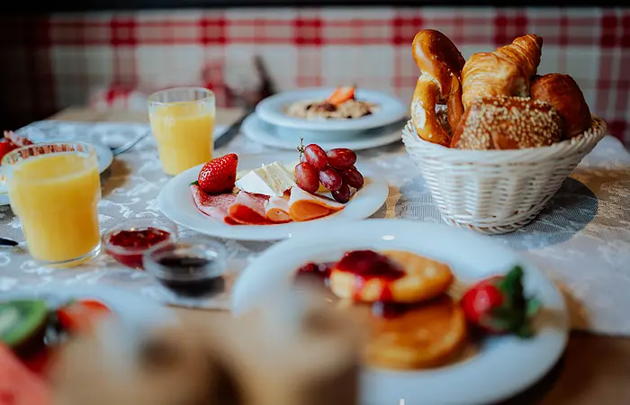 Der Frühstückstisch ist mit Orangensaft, Marmeladen, Pancakes und einem gut gefüllten Brötchenkorb gedeckt.