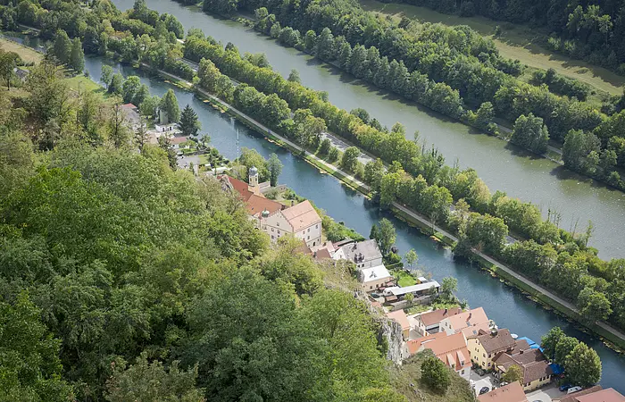 Wir blicken von oben auf Essing am Main-Donau-Kanal. Die Ortschaft ist von Wald, Felsen und auf der anderen Seite vom Kanal umgeben.