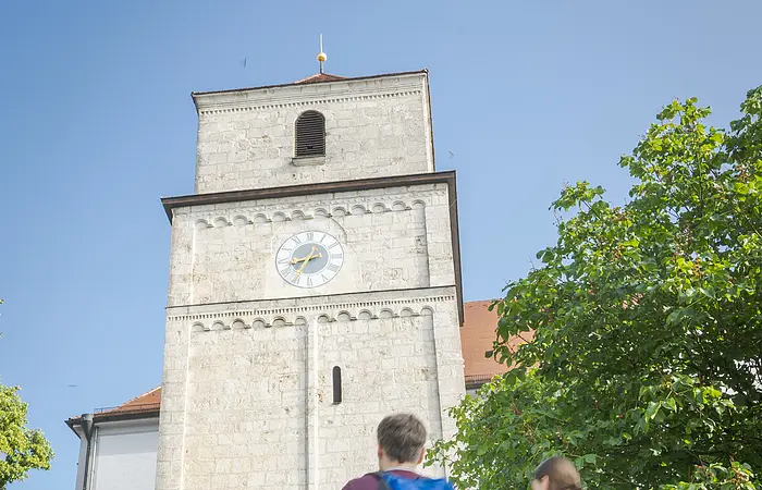 Zwei Personen mit Rucksäcken gehen auf einen alten Turm mit Uhr zu, umgeben von Bäumen.
