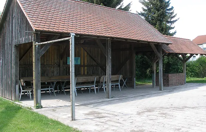 Überdachter Holzpavillon mit Tischen und Bänken auf gepflastertem Boden neben Rasenfläche und Bäumen.