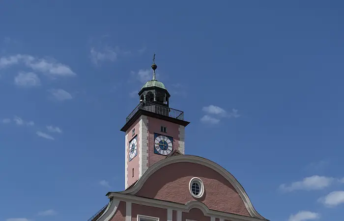 Das Bild zeigt ein prächtiges vierstöckiges Haus mit einem geschwundenen Giebel. Darüber thront im Hintergrund ein Glockenturm mit Ziffernblätter und einem Kupferdach sowie einem Balkon, der um den gesamten Turm führt. Das Rathaus und der Turm sind rotbraun gestrichen. Der Eingang in Form eines zweiflügeligen Holztors befindet sich in der Mitte der Vorderseite, direkt darüber ist ein Balkon zu erkennen, der durch zwei Säulen links und rechts des Eingangstores gestützt wird.