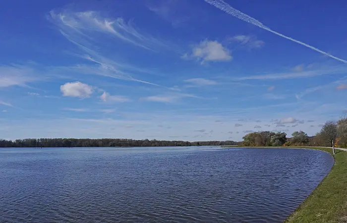 Uferweg entlang eines großen Gewässers unter blauem Himmel mit wenigen Wolken und Baumreihe am Horizont.