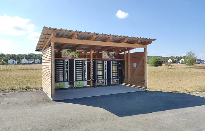 Auf einer Asphaltfläche befindet sich ein Holzbau im Format eines Carports, nach vorne offen. Darin stehen vier Automaten. Im Hintergrund sieht man ein reifes Getreidefeld, dahinter ein Streifen Bäume und blauer Himmel.