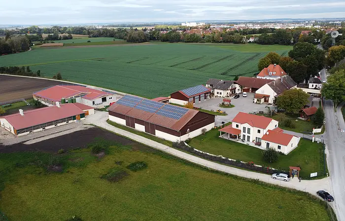 Bild von oben mit der gesamten Hofanlage. 2 Scheunen und mehrere Hauser mit weißen Wänden und roten Dächern. Ein Weg führt zu der Hofstelle. Umgeben ist die Anlage von grünen Feldern und Wiesen. Am Rand der Häuser stehen Bäume.