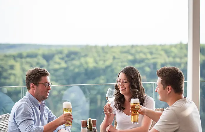 Zwei Männer und eine Frau, alle mit vollen Getränken in der Hand, prosten sich zu. Hinter ihnen hat man einen wunderschönen Blick ins Tal und auf die Wälder.
