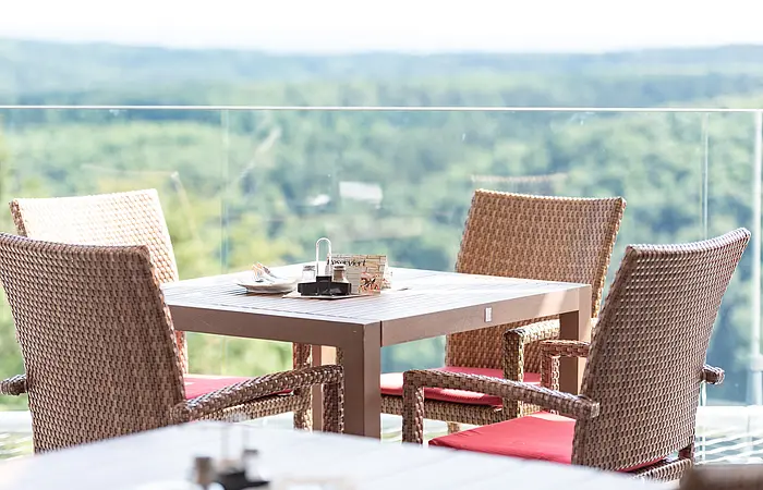Ein Tisch mit vier Rattanstühlen mit roten Kissen. Dahinter eine Glasbrüstung und der wunderschöne Blick ins Tal und den grünen Wäldern.