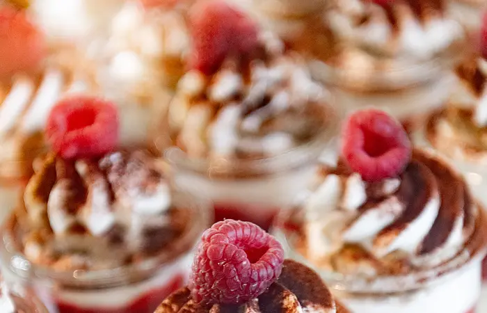 Dessertgläschen mit weiß-roter Füllung, kakaobestäubtem Häubchen und Himbeere garniert, stehen aufgereiht bereit serviert zu werden.