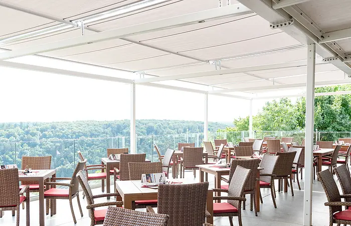 Die Terrasse mit Holztischen und Stühlen ist komplett überdacht. Auf allen Tischen sind rote Tischdeckchen mit Besteck und Speisekarte zu erahnen. Die Terrasse gewährt einen wunderschönen Blick ins Tal.