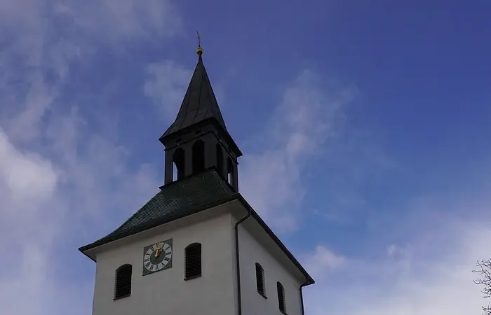 Kirchturm mit Uhr und spitzem Dach vor blauem Himmel, im Vordergrund weiße Mauer mit Torbogen.