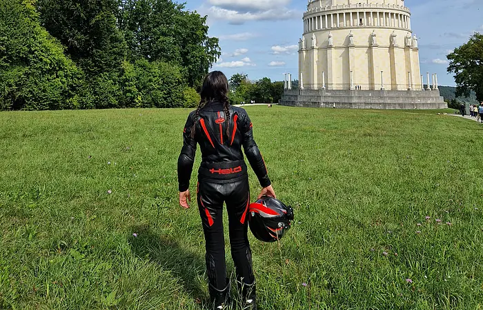 Person in schwarzer Motorradkleidung mit Helm in der Hand steht auf Wiese vor rundem Gebäude mit Säulen und Statuen.