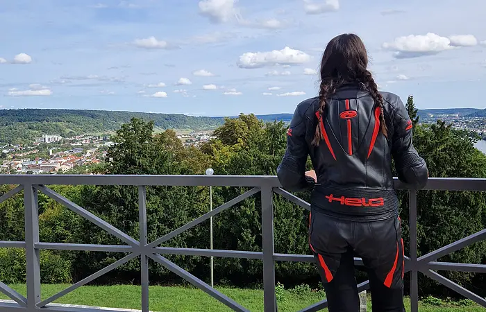 Person in schwarzer Motorradkleidung mit roten Akzenten blickt von Balkon auf grüne Landschaft und Stadt, Helm liegt am Boden.