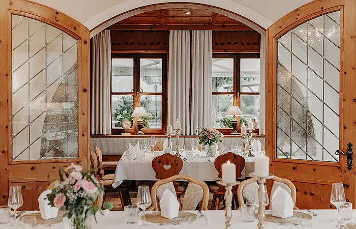 Ein schöner gedeckter Tisch mit weißer Tischdecke, roten Blumen und Kerzen. Dahinter stehen die hölzernen Türen auf und lassen uns einen Blick in den nächsten Gastraum erhaschen.