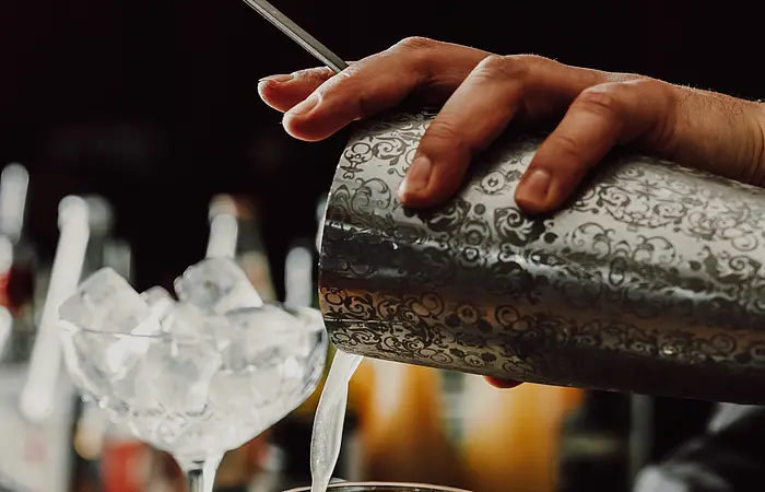Zwei Gläser mit Eiswürfel stehen auf dem Tresen. Der Bartender gießt in das vordere den Cocktail in das Glas. Im Hintergrund sind die verschiedenen Flaschen zu erkennen.