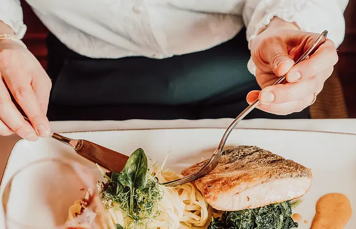 Mit Blick von oben auf einen gefüllten TEller mit grüner Beilage, rötlicher Soße und einem Stück Fleisch. Im Vordergrund steht ein Weinglas mit Rotwein gefüllt. Der Oberkörper des Gastes mit weißem Hemd ist zu sehen. Er hat sich soeben ein Stück abgeschnitten.
